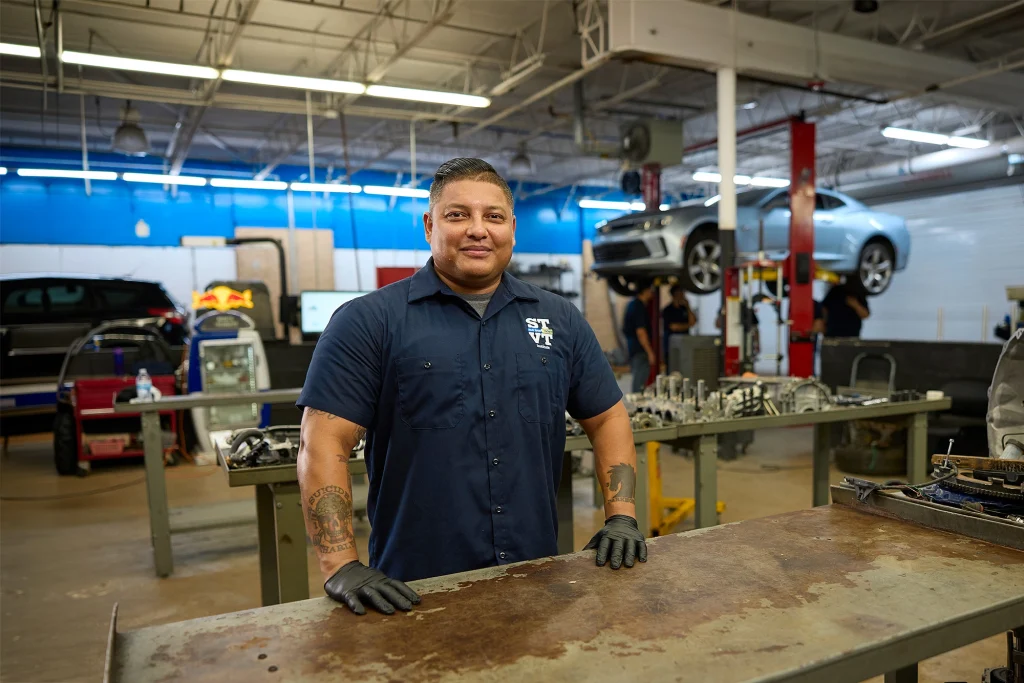 STVT auto technician in a navy work shirt standing at a workbench in an automotive shop with vehicles on lifts in the background.